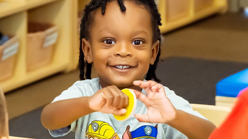 A child holds up a puzzle piece, smiling, at the camera