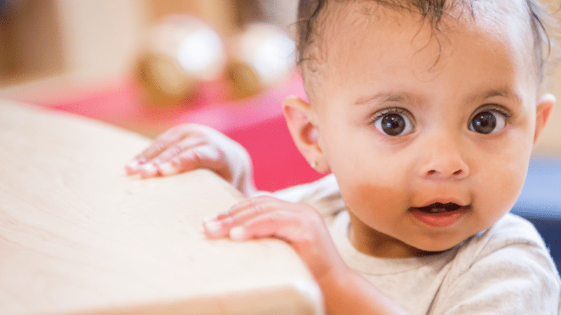 A young child looking directly at the camera holding onto a surface for support