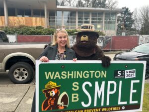 Photo of Notes From Olympia author Erica Hallock with Smokey the Bear holding a large sample license plate advertising the design of proposed Specialty License Plate featuring Smokey the Bear.